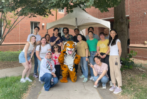 a group of 16 people pose for a photo around a person wearing a tiger mascot suit